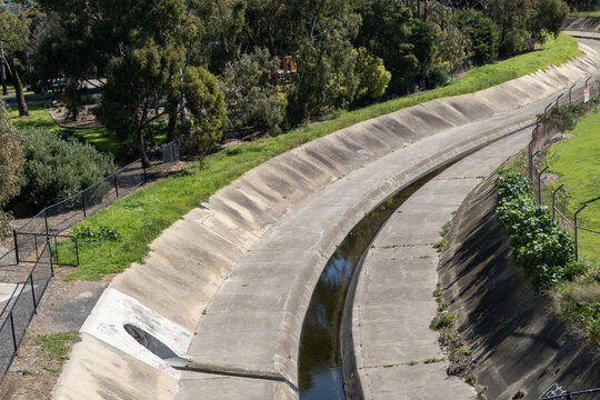 Concrete lined drainage channel of Stony Creek curving past chain link security boundary fencing and parkland trees in Yarrville, Melbourne, Victoria, Australia