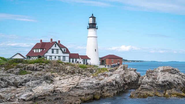 Portland Head Light timelapse standing atop rocky coastal cliffs at Cape Elizabeth, Maine, with visitors walking around the grounds and ocean waves churning along Casco Bay.