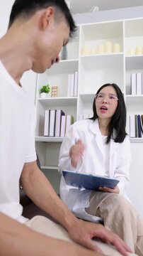 Professional Asian female physician talking to a male patient, listening to his problems and providing consultation and diagnosis in a clinic.
