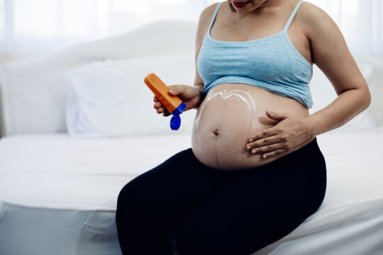Pregnant woman applying lotion in heart shape to belly while sitting on bed, showing maternity skincare, moisturizer, pregnancy care, hydration, bonding and prenatal wellness