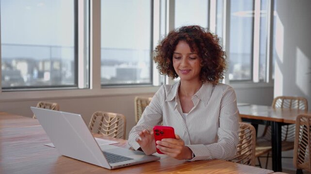 Young latin business woman using smartphone phone at laptop in sunlit coworking office, static shot. Smiling hispanic specialist checks online message, browsing mobile banking application at company