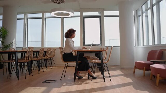 Wide shot of hispanic business woman sitting alone at table with laptop by panoramic office window. Young latin professional working online in empty modern coworking space, checking company data