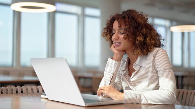 Young latin business woman typing on laptop in modern coworking office, thinking with hand on chin. Smiling hispanic professional works online, checking company financial data and project report