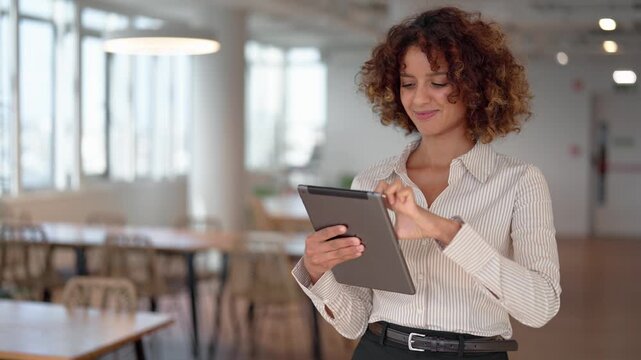 Latin business woman scrolling digital tablet in sunlit coworking office, static shot, smiling at screen. Young hispanic specialist checking online market data, browsing fintech application at work