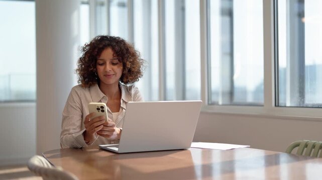Latin business woman working on laptop with paper document in modern coworking office at long table. Young hispanic accountant checks financial report online, planning company project and budget