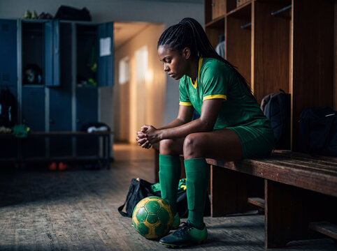Tears of Defeat: A lone female soccer player, cloaked in disappointment, sits dejectedly in a locker room after a tough game, her football resting by her side.