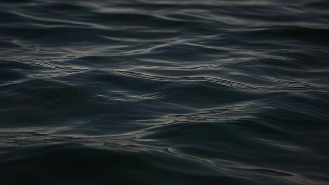 Dark sea surface with gentle wave patterns, reflective ripples, silver water reflection, moody abstract texture, tranquil natural background, coastal imagery, ocean pattern design