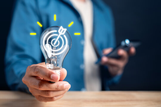 Business idea innovation and strategy goal objective success. Man hand holding lightbulb with target bullseye arrow and smartphone device.
