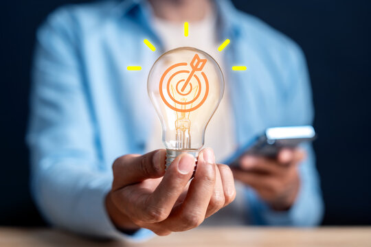 Business idea innovation and strategy goal objective success. Man hand holding lightbulb with target bullseye arrow and smartphone device.