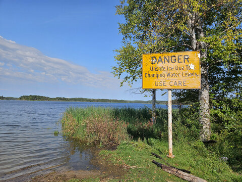 Humorous yellow sign warning of the dangers of thin ice  is in contradiction to the lake of calm water it is standing next to during the summer.