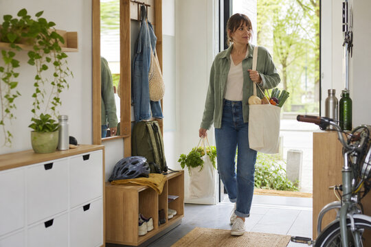 Woman returning home with reusable grocery bags full of fresh vegetables beside a bicycle in an eco friendly plant filled entryway for a greener future at home