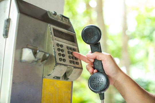 Selective focus to hand holding a black receiver and dialing a number on an old public payphone booth outdoors, set against a blurred green natural background.