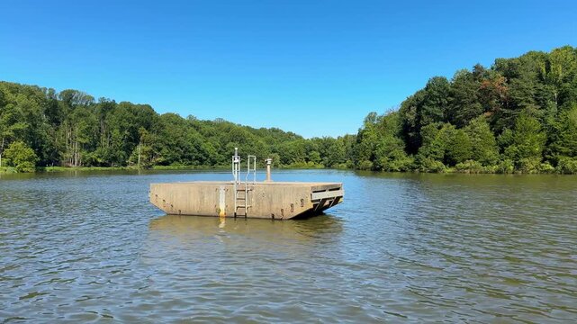 Concrete dam spillway riser tower on a serene lake under bright natural daylight. Horizontal video showcasing peaceful nature aesthetics, blue sky, and a dense green forest backdrop.