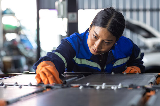 Female electric vehicle technician inspecting EV battery pack in automotive workshop, showing high-voltage battery maintenance, skilled engineering, clean mobility and electric car technology.