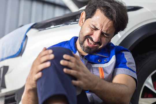Automotive mechanic holding painful knee beside vehicle in workshop, workplace injury, occupational safety, physical strain, accident risk and health concerns in car repair work.