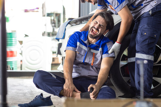 Injured automotive mechanic holding his ankle while coworker assists him on workshop floor, showing workplace accident, occupational safety, worker injury, emergency support and accident risk.