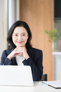 A female office worker working on a laptop; a businesswoman working with a smile in the office, resting her hand on her chin, looking directly at the camera while doing desk work (portrait orientation
