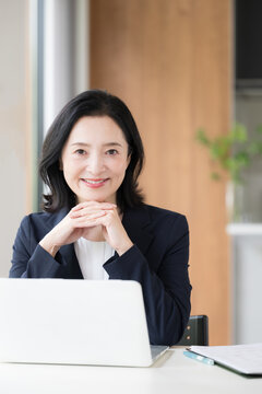 ノートパソコンで仕事をする女性会社員、オフィスで笑顔で働く顎に手を当てるビジネスウーマンのデスクワークカメラ目線縦