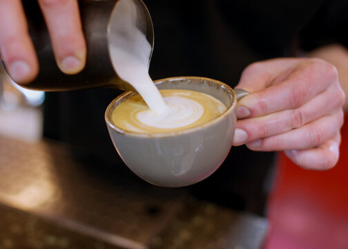 Latte art. Pouring foamed milk. Barista hand adding frothing creamy component from pitcher to beverage garnished coffee in ceramic cup.