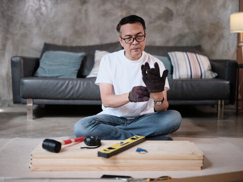 One Asian mature man wearing protective gloves prepares DIY tools and flat-pack furniture for assembly, builds for home improvement, wooden craft install hobby workshop, handyman active lifestyle.