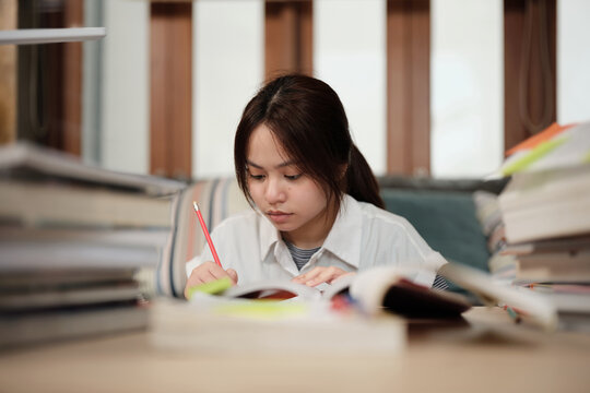 Asian teenage girl concentrates on pile of books, focuses on learning at home with pile of textbooks, reading and writing, self-study for school, Homeschooling, university education lifestyle.
