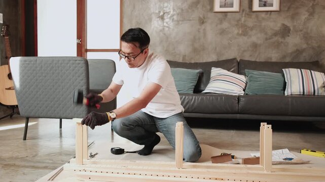Asian mature man self-assembling flat-pack furniture with DIY tools, nailing joint parts with rubber mallet, home improvement, craft installation, hobby wooden workshop, handyman active lifestyle.