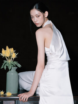 Studio portrait of Chinese woman in white satin dress beside flower vase, gazing over shoulder with calm expression against black background
