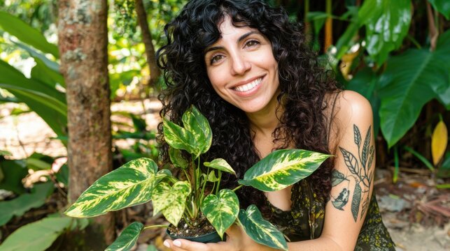 A smiling woman with curly hair holds a variegated potted plant outdoors, surrounded by lush greenery, showcasing a joyful connection with nature.