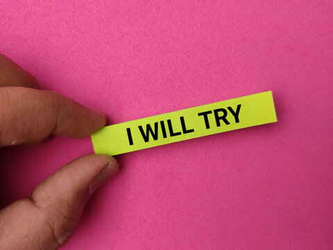 A hand holds a bright yellow strip printed with the text I WILL TRY against a vibrant pink background