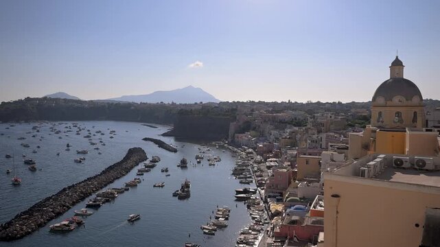 Colorful Marina Corricella waterfront and Santa Maria delle Grazie Incoronata rise above a busy harbor filled with small boats on a clear sunny day.