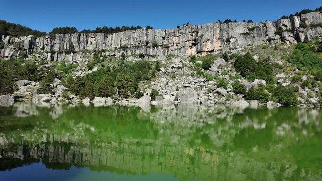 Handheld wide angle footage of Laguna de Urbi&oacute;n's emerald water, steep rocky cliffs, pine forest, and vivid sky, captured on a bright day.