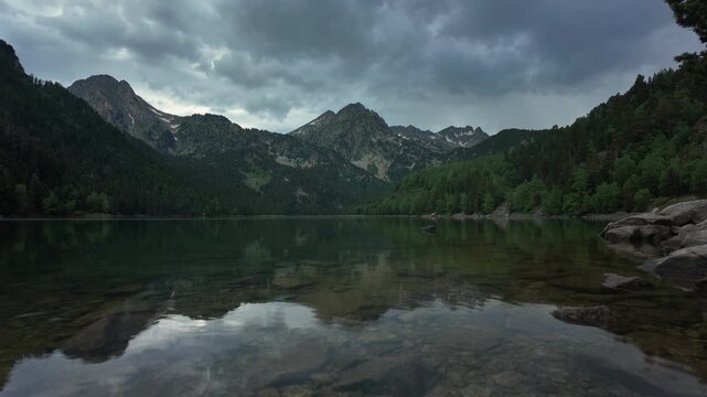 Dramatic timelapse captures moody storm clouds gathering above Estany de Sant Maurici alpine lake in Parc Nacional d'Aig&uuml;estortes, Pyrenees, with dark reflections on still waters.