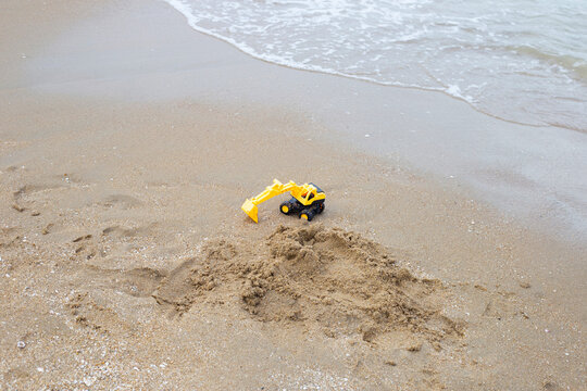 A yellow toy excavator digging in the sand on a tropical beach with sea waves.