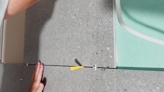 Close-up of a worker pressing a large gray wall tile firmly into place, aligning spacers during interior renovation and professional ceramic wall installation.