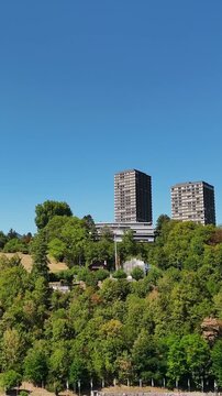 Tilt-up drone shot from the turquoise Rhine near Rhine Falls to wooded hills and modern high-rise towers in Schaffhausen, Switzerland, under a clear blue summer sky.