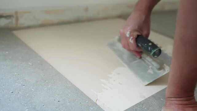 Close-up of a worker smoothing fresh tile adhesive across a floor substrate with a trowel, preparing an even bonding layer during interior flooring installation.