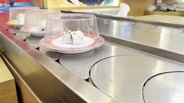 Fresh sushi on covered plates moving on a conveyor belt system at a kaiten-sushi restaurant in Japan. Traditional Japanese dining experience, maki rolls under plastic domes.