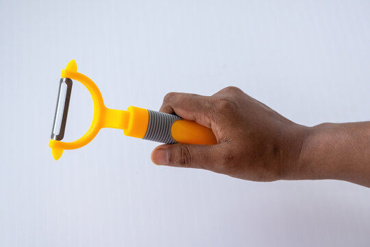 Person holding a bright yellow vegetable peeler with stainless steel blade, modern handheld kitchen tool isolated over white studio background.