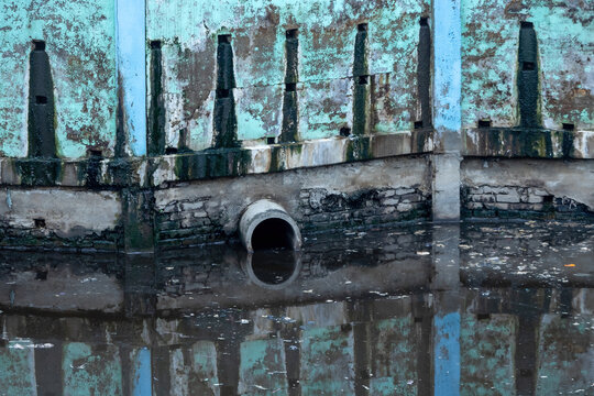 Industrial concrete drainage pipe protruding over stagnant dirty canal water with floating garbage debris.