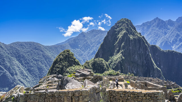 The famous ancient Machu Picchu. People are walking among the half‑ruined masonry walls, looking at the landmark. Huayna Picchu and the Andes mountain range against a blue sky and clouds. Peru. Cusco.