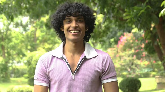 Young indian Gen Z man with curly hair smiling looking at camera outdoors in summer park, concepts of joy, happiness, and a positive lifestyle.