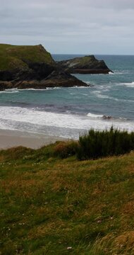 Sandy beach and rugged cliffs in Cornwall, England, UK, with breaking waves and coastal grass beneath a cloudy afternoon sky. Vertical handheld footage.
