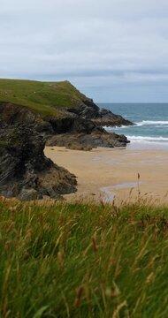 Sandy beach and rugged cliffs in Cornwall, England, UK, with breaking waves and coastal grass beneath a cloudy afternoon sky. Vertical handheld footage.