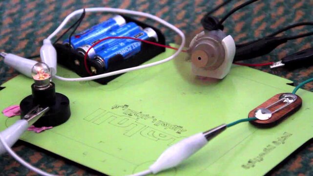 A simple educational electrical circuit featuring a battery pack, a glowing light bulb, and a small motor connected by wires on a green board.