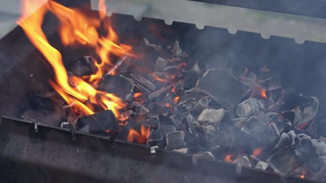 Close-up of hot charcoal briquettes burning with bright orange flames and smoke inside a metal barbecue grill, getting ready for cooking outdoors during a picnic or backyard party