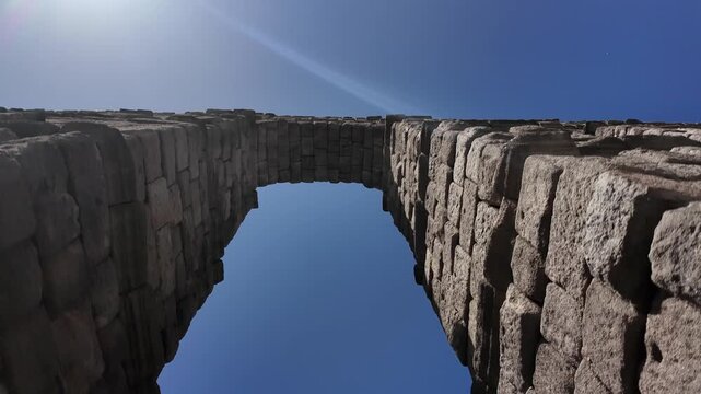 Dynamic handheld view under Segovia's ancient stone aqueduct as swifts dart past massive arches against vibrant blue sky, highlighting iconic Roman architecture in crisp natural daylight.