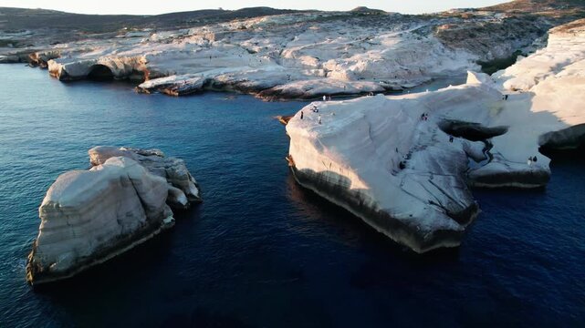 Drone footage over Sarakiniko, Milos. Stunning white volcanic formations curve gracefully over the sea under a clear blue sky. Showcasing tourists exploring the beautiful scenery