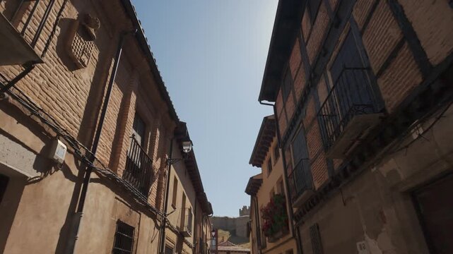 Handheld camera glides under sunlit, narrow stone streets between rustic brick facades and iron balconies in Berlanga de Duero Village