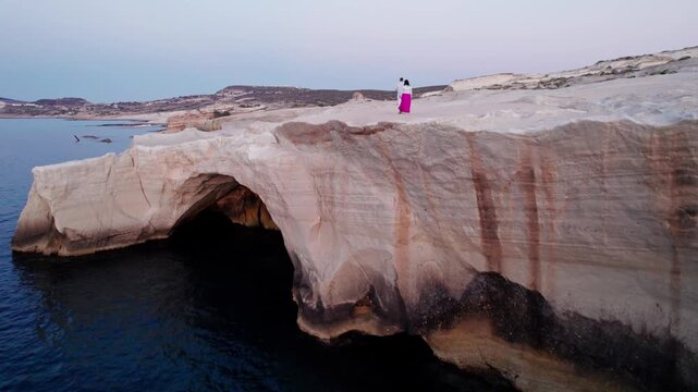 Flyover aerial view of Sarakiniko&rsquo;s white volcanic coastline and sea arch in Milos, Greece, with tourists atop the cliff.