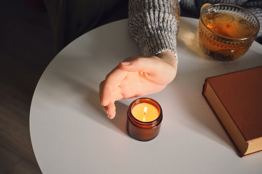 Female hand near burning candle on table with cup of tea and book
